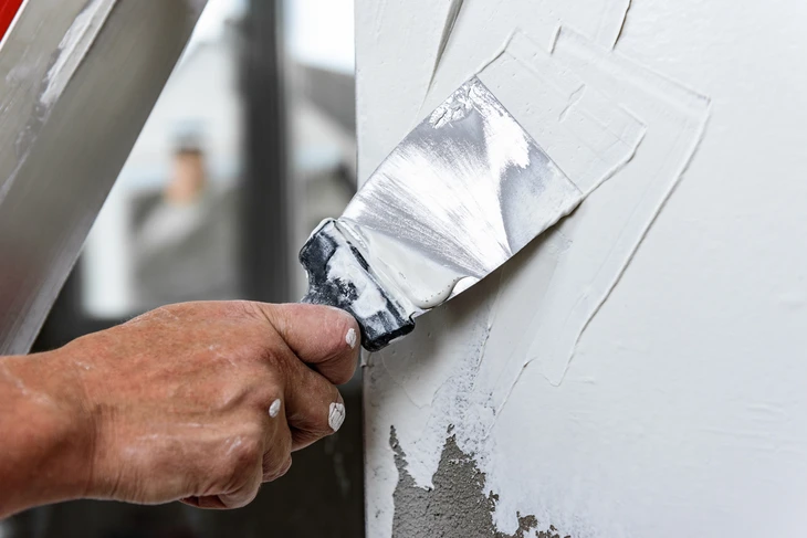 Une main tenant un couteau à enduire applique un enduit de lissage blanc sur un mur intérieur, à la jonction entre une surface grise et blanche